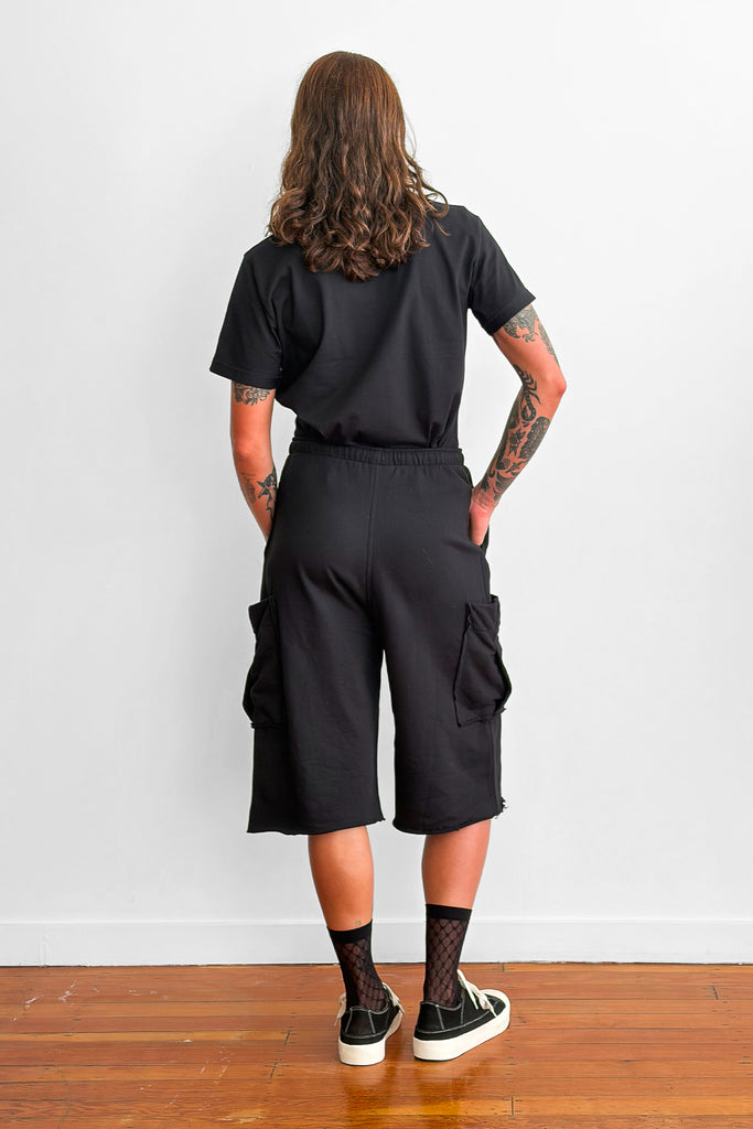 Back view of a model wearing a black t-shirt tucked into oversized, long black shorts