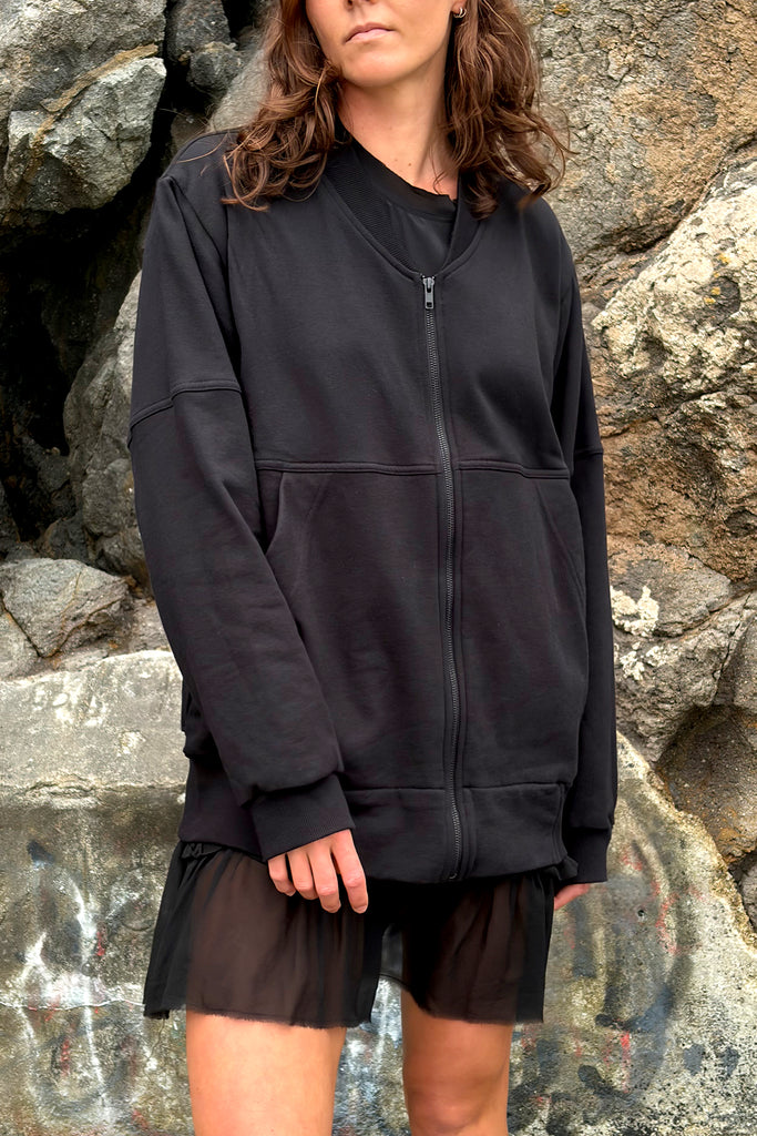 Model wearing an oversized black zip sweater over a mini black dress. Standing infront of a rocky wall